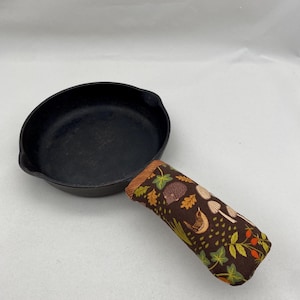 May include: A black cast iron skillet with a brown fabric handle featuring a woodland pattern of birds, hedgehogs, mushrooms, and leaves.