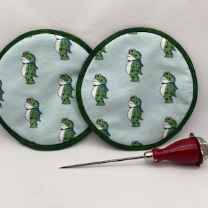 May include: Two round, light blue fabric coasters with a green border feature a repeating pattern of green frogs wearing blue scarves. A red and silver awl with a long, silver needle is also in the image.