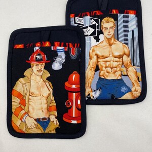 May include: Two black pot holders with a firefighter theme. One features a shirtless firefighter in a tan jacket and red helmet. The other depicts a muscular man in blue shorts with a dog, and a city backdrop. The pot holders have black trim.