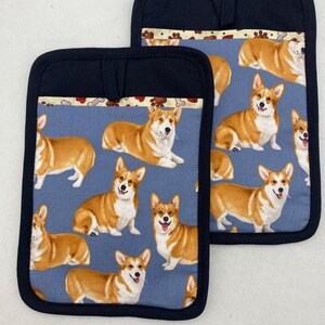May include: Two rectangular pot holders featuring a repeating pattern of brown and white Corgi dogs on a blue background. Each pot holder has a solid black fabric border and a small fabric loop at the top center for hanging, with a decorative trim.