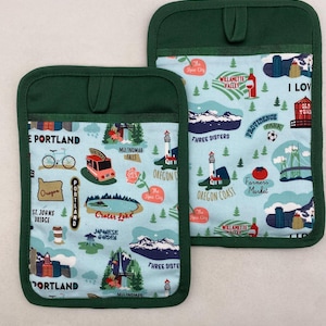 May include: Two matching pot holders with a light blue background featuring Oregon landmarks like Crater Lake and Multnomah Falls. The pot holders have a dark green trim and a loop for hanging.