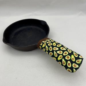 Puede incluir: Sartén de hierro fundido negro con una cubierta de mango de tela con estampado de aguacate marrón y negro.