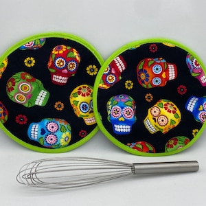 May include: Two round black and green hot pads with a colourful sugar skull pattern. The hot pads are on a white surface with a silver whisk.