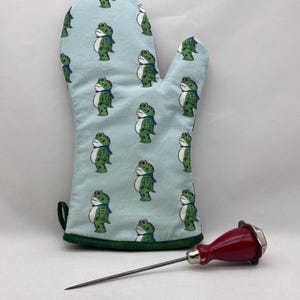 Puede incluir: Un guante de cocina azul claro con un ribete verde y un patrón repetido de ranas verdes de dibujos animados con bufandas azules. Un termómetro para carne con mango rojo y punta plateada también está en la imagen.