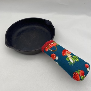 Op de afbeelding: Een zwarte gietijzeren koekenpan met een teal en rode stoffen handvat met een paddenstoelmotief.