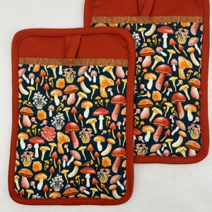 Op de afbeelding: Twee bijpassende pannenlappen met een donkerblauwe achtergrond en een kleurrijk paddenstoelenpatroon. De pannenlappen hebben een roestkleurige rand en een lus om op te hangen. Het paddenstoelontwerp bevat rode, oranje, gele en witte paddenstoelen.