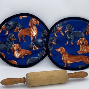 May include: Two round blue pot holders with a black trim, featuring a repeating pattern of brown and black dachshund dogs. A wooden rolling pin rests below the pot holders.