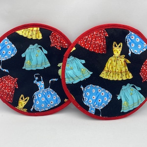 Op de afbeelding: Twee ronde stoffen pannenlappen met een zwarte achtergrond en een patroon van vintage jurken in rood, geel en blauw. De randen zijn afgewerkt met rode stof.