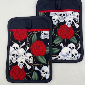 May include: Two black pot holders with a repeating pattern of white skulls, red roses, and green leaves. The pot holders have a black border and a red stripe near the top. The design is set against a black background.