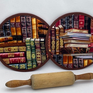 May include: Two round brown pot holders with a book print design. One pot holder features a stack of books and the other features a bookshelf full of books. A wooden rolling pin is in the foreground.