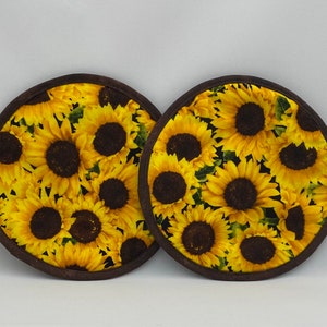 Può includere: Due sottopentola rotondi di colore marrone e nero con un motivo a girasole. I sottopentola sono realizzati in tessuto e hanno un bordo marrone.