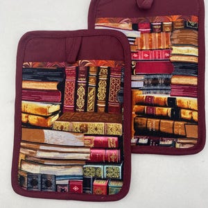 May include: A pair of burgundy pot holders with a book stack design. The pot holders are made of a thick, quilted fabric and have a loop for hanging.