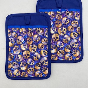 Könnte beinhalten: Zwei rechteckige Topflappen mit einem sich wiederholenden Muster aus goldbraunen Totenköpfen vor einem lebendigen, von Galaxien inspirierten Hintergrund in Violett und Blau. Jeder Topflappen hat eine dunkelblaue Stoffkante und eine passende Aufhängeschlaufe.