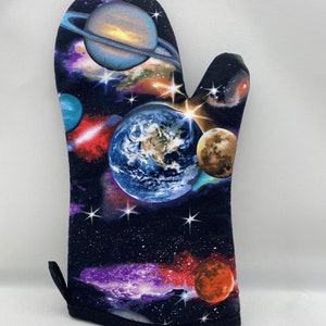 Pode incluir: Uma luva de forno preta com um design temático espacial. O design apresenta planetas, estrelas e galáxias em tons de azul, laranja, roxo e branco. Possui uma alça para pendurar.