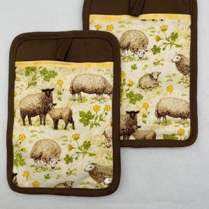 May include: Two brown and beige pot holders with a sheep pattern. The pot holders have a brown border and a loop for hanging. The fabric features sheep, yellow flowers, and green foliage on a beige background.