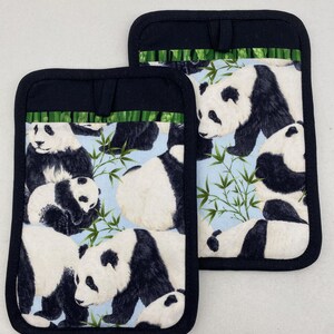 Puede incluir: Dos manoplas para horno con osos panda en blanco y negro. Las manoplas tienen un borde negro y un lazo para colgar. Los osos panda están sobre un fondo azul claro con tallos de bambú.