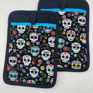May include: Two black oven mitts with a colorful sugar skull pattern and a turquoise trim.