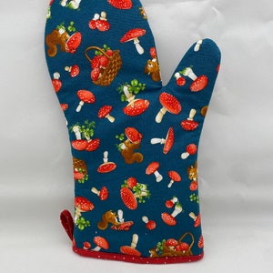 Puede incluir: Un guante de horno azul con un patrón de setas rojas y blancas, hojas verdes y ardillas marrones. El guante tiene un ribete rojo con lunares blancos.