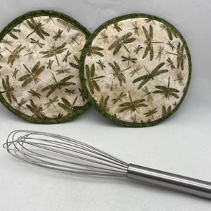 May include: Two round, dragonfly-patterned pot holders with a green border sit above a stainless steel whisk. The pot holders feature a cream background with brown dragonflies. The whisk has a silver handle.