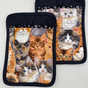 May include: Two black oven mitts with a pattern of colorful kittens. The mitts have a pocket on the back.