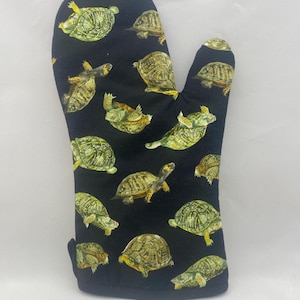 Peut inclure: Un gant de cuisine noir avec un motif répété de tortues détaillées dans des tons de vert, de marron et de jaune. Le gant a un pouce et une boucle pour suspendre. Un accessoire de cuisine pour cuisiner et pâtisser.