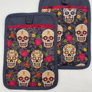 May include: Two rectangular oven mitts with a dark grey border and a loop for hanging. The main fabric features a pattern of colourful sugar skulls, red roses, and yellow flowers on a dark grey background. The top edge has a red ribbon.