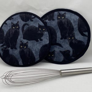 May include: A set of two black cat-themed pot holders. The pot holders are round and have a blue background with black cats printed on them. The cats are in various poses, including sitting, standing, and lying down.