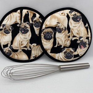 May include: Two black and white round pot holders with a pattern of pugs. The pugs are beige and brown with black noses and eyes. The pot holders are on a white surface with a silver whisk.