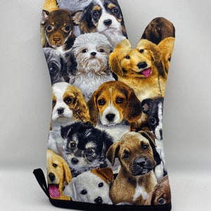 May include: A single oven mitt featuring a collage of various dog breeds. The mitt is covered with images of dogs in different colors and sizes, with a black trim and a black hanging loop.