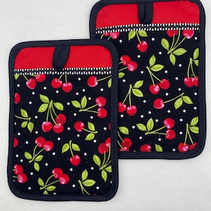 May include: Two black pot holders with a red cherry pattern and a red top border. The cherry pattern includes red cherries, green leaves, and white polka dots. Each pot holder has a black loop for hanging.