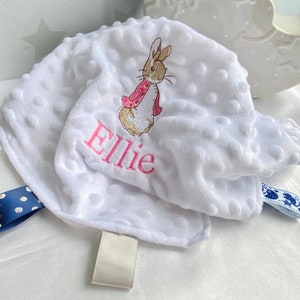 Może przedstawiać: Biały kocyk dla dziecka z haftowanym różowym króliczkiem i imieniem "Ellie" w kolorze różowym. Kocyk ma przymocowaną niebiesko-białą wstążkę.