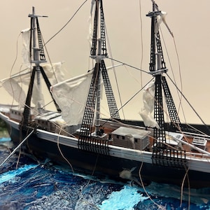 May include: A detailed model ship with black hull, white sails, and intricate rigging. The ship is set in a sea of blue and white waves, with a rocky shoreline in the background. The model is a miniature replica of a sailing vessel.