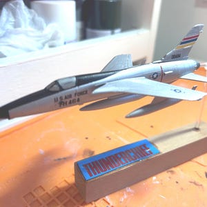 May include: A silver model airplane with black accents and the text "U.S. AIR FORCE" and "TH 464" on the side. The model is mounted on a wooden base with the word "THUNDERCHIEF" on a blue label.