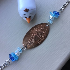 May include: A silver chain bracelet with a copper-colored coin charm featuring a princess silhouette and the word "Disneyland". The bracelet is decorated with blue and clear glass beads.