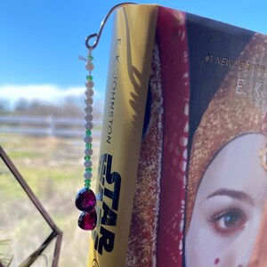 May include: A bookmark with a silver wire, white and green beads, and two purple gemstone charms. The bookmark is attached to a book titled "Star Queen's Shadow" by E.K. Johnston.