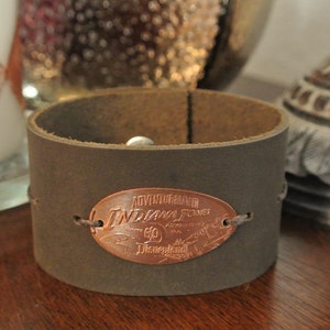 May include: A brown leather cuff bracelet with a copper-colored oval charm that reads "Adventure Bound Indiana Jones", with a small "60" and "Discovered" text.