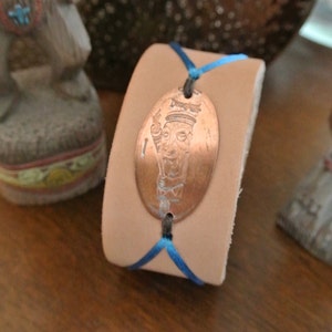 May include: A tan leather cuff bracelet with a copper oval pendant. The pendant features an engraved image and is attached to the bracelet with blue cord. The bracelet is displayed on a wooden surface.