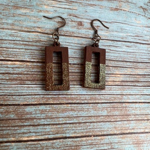 May include: A pair of brown wooden earrings with gold glitter accents. The earrings are rectangular in shape with a square cutout in the center. The earrings have a hook closure.
