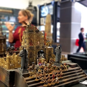 May include: A detailed miniature model of a medieval city, featuring a large, ornate building with a pointed archway, surrounded by smaller buildings and figures. The model is painted in gold and silver tones, with intricate details.