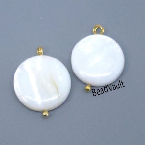 May include: Two white, round pendants with a pearlescent sheen. Each pendant has a gold-colored loop at the top and a small gold bead at the bottom. The pendants are set against a gray background. The text "BeadVault" is visible.
