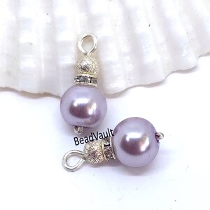 May include: Two lavender pearl charms featuring a silver-toned metal loop at the top, a textured silver bead, and a small ring of clear rhinestones. The smooth, round lavender pearls are positioned vertically and horizontally on a white surface.