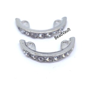 May include: Two silver-toned, curved jewelry findings with clear rhinestones. Each finding has a series of small, round rhinestones set along its curved surface. The text "BeadVault" is visible on the image.