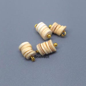 May include: Three jewelry charms, each consisting of a stack of small, circular, off-white shell beads threaded onto a gold-toned metal pin with a loop at one end and a small gold-toned ball at the other. The text BeadVault is visible.