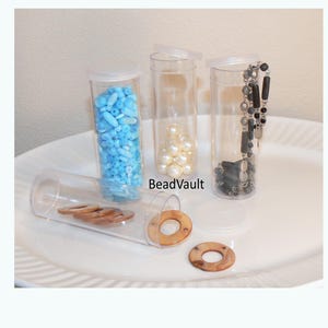 May include: Four clear cylindrical plastic storage tubes with white lids. One tube contains light blue beads, one holds white pearls, one contains a black and silver beaded necklace, and one lying horizontally holds brown wooden rings. BeadVault text.