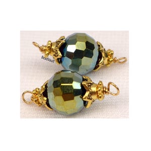 Può includere: Due perline di vetro sfaccettate iridescenti con dettagli in metallo color oro. Le perline hanno una forma sferica e una superficie sfaccettata che riflette sfumature di verde e blu. La lavorazione del metallo presenta un design a forma di stella e un anello per l'infilatura.