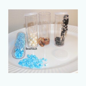 May include: Three clear cylindrical plastic containers with lids, one lying on its side filled with light blue beads, another containing white spherical beads, and a third holding a black beaded chain. The text BeadVault is visible.