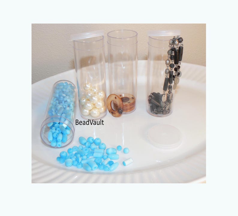 May include: Three clear cylindrical plastic containers with lids, one lying on its side filled with light blue beads, another containing white spherical beads, and a third holding a black beaded chain. The text BeadVault is visible.