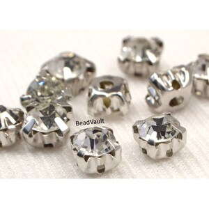 May include: A close-up of ten clear crystal rhinestones with silver settings. The rhinestones are arranged in a cluster on a white fabric background. BeadVault is visible in the image.