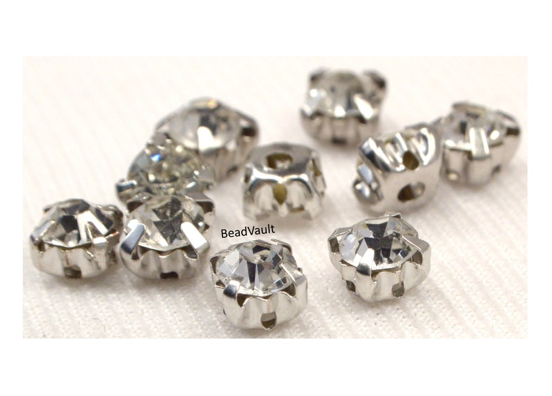 May include: A close-up of ten clear crystal rhinestones with silver settings. The rhinestones are arranged in a cluster on a white fabric background. BeadVault is visible in the image.