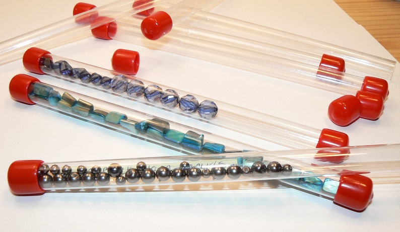 May include: Six clear plastic tubes with red caps, filled with various small beads and metal balls. The tubes are arranged in a row on a white surface.